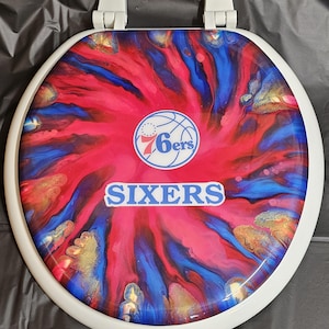 May include: A white toilet seat with a red, blue, and gold abstract swirl design. The Philadelphia 76ers logo is in the center with the word "SIXERS" below it.
