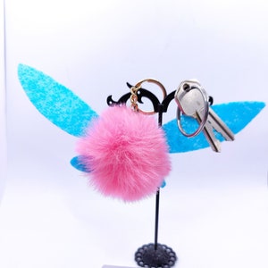 May include: Pink fluffy pom-pom keychain with a silver key and a gold chain attached to a silver key ring. The keychain is displayed on a black stand with blue wings. The tag attached to the stand reads "dark dainty" and "darkdainty.com".