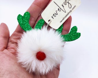 Op de afbeelding: Witte, pluche pompon rendier ornament met groene glittergewei en een rode pompon neus. De ornament heeft een zilveren ring om op te hangen en een label met de tekst "revelrose revelroseshop.com".