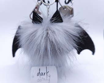 May include: A white fluffy pom-pom keychain with black felt bat wings. The keychain has a silver metal ring and a silver metal tag with the text "dark & dainty" and the website "darkdainty.com".