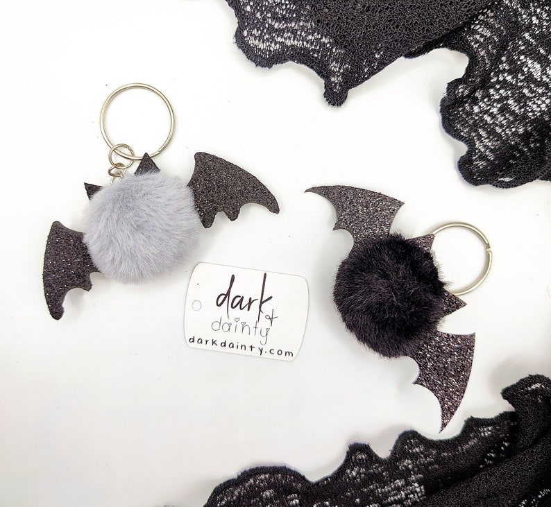 Op de afbeelding: Twee vleermuisvormige sleutelhangers met glinsterende zwarte vleugels en pluizige pompons, &eacute;&eacute;n grijs en &eacute;&eacute;n zwart, met zilveren metalen ringen. Een wit label toont de tekst dark dainty en darkdainty.com in zwarte letters.