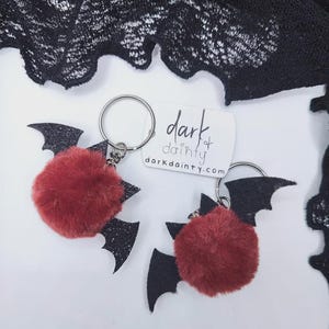 Op de afbeelding: Twee sleutelhangers met pluizige, ronde, donkerrode pompons bevestigd aan zwarte glitterende vleermuisvormige vleugels. Elke sleutelhanger heeft een zilveren metalen ring. Een wit rechthoekig label bevat de tekst dark & dainty en darkdainty.com.