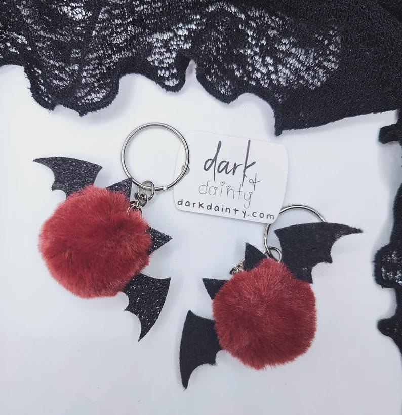 Op de afbeelding: Twee sleutelhangers met pluizige, ronde, donkerrode pompons bevestigd aan zwarte glitterende vleermuisvormige vleugels. Elke sleutelhanger heeft een zilveren metalen ring. Een wit rechthoekig label bevat de tekst dark & dainty en darkdainty.com.