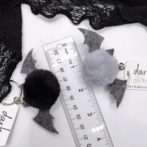Op de afbeelding: Twee vleermuisvormige sleutelhangers met pluizige pompons, &eacute;&eacute;n zwart en &eacute;&eacute;n grijs, met viltvleugels en metalen ringen. Een doorzichtige liniaal toont een breedte van ongeveer 6,5 cm. Witte labels met de tekst darkdainty zijn bevestigd.