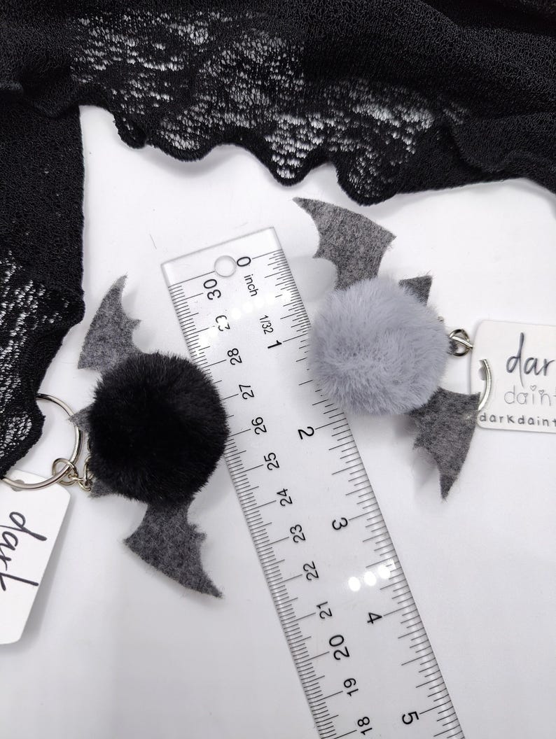 Op de afbeelding: Twee vleermuisvormige sleutelhangers met pluizige pompons, &eacute;&eacute;n zwart en &eacute;&eacute;n grijs, met viltvleugels en metalen ringen. Een doorzichtige liniaal toont een breedte van ongeveer 6,5 cm. Witte labels met de tekst darkdainty zijn bevestigd.