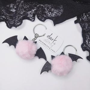 Op de afbeelding: Twee vleermuis-sleutelhangers met pluizige lichtroze pompons bevestigd aan zwarte glitterende viltvleugels en zilverkleurige metalen ringen. Een wit rechthoekig label bevat de tekst dark dainty en darkdainty.com in zwarte inkt.