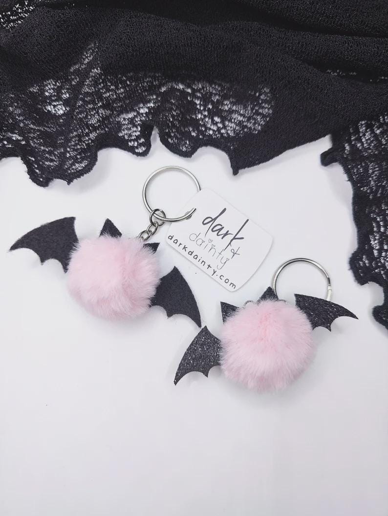 Op de afbeelding: Twee vleermuis-sleutelhangers met pluizige lichtroze pompons bevestigd aan zwarte glitterende viltvleugels en zilverkleurige metalen ringen. Een wit rechthoekig label bevat de tekst dark dainty en darkdainty.com in zwarte inkt.