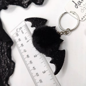 Op de afbeelding: Een zwarte glitterende vleermuisvormige sleutelhanger met een centrale zwarte imitatiebont pompon, bevestigd aan een zilveren metalen ring. Een liniaal toont aan dat de vleermuis ongeveer 7,6 cm breed is. Een wit label bevat de tekst dark daisy.