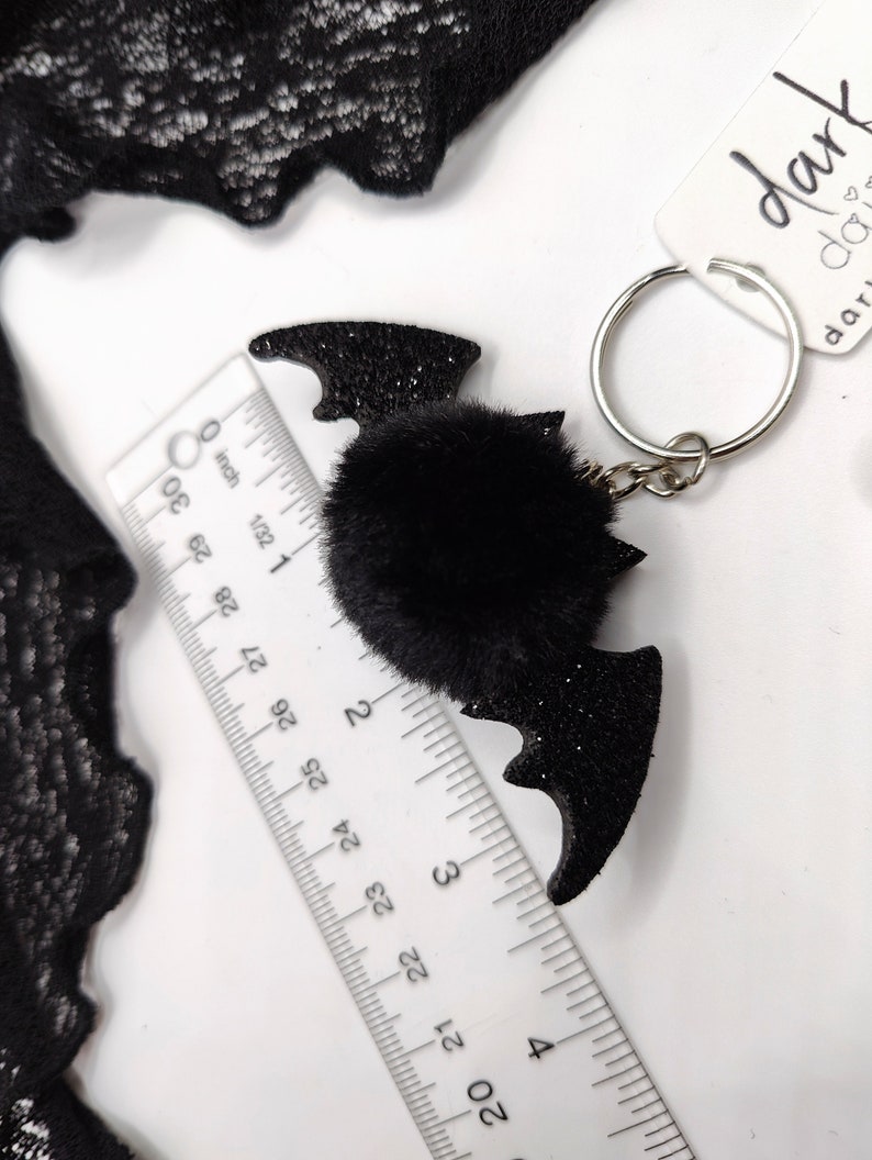 Op de afbeelding: Een zwarte glitterende vleermuisvormige sleutelhanger met een centrale zwarte imitatiebont pompon, bevestigd aan een zilveren metalen ring. Een liniaal toont aan dat de vleermuis ongeveer 7,6 cm breed is. Een wit label bevat de tekst dark daisy.