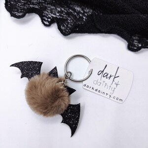 Op de afbeelding: Een sleutelhanger met een bruine imitatiebont pompon bevestigd aan zwarte glitterende vleermuisvleugels. Een zilveren metalen ring verbindt de vleermuis met een wit rechthoekig label waarop de tekst dark dainty en het webadres darkdainty.com staat.