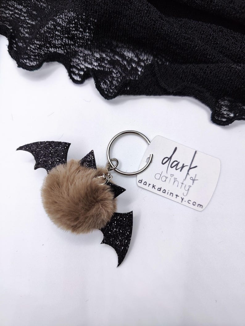 Op de afbeelding: Een sleutelhanger met een bruine imitatiebont pompon bevestigd aan zwarte glitterende vleermuisvleugels. Een zilveren metalen ring verbindt de vleermuis met een wit rechthoekig label waarop de tekst dark dainty en het webadres darkdainty.com staat.