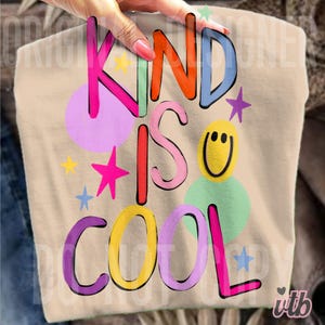May include: A beige t-shirt with the words "KIND IS COOL" in colorful bubble letters. The design includes stars and a smiling face. The shirt is held up by a person.