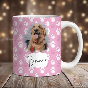 May include: A pink ceramic mug with a white paw print pattern. A photo of a golden retriever dog is in the center of the mug with the name "Bonnie" written in a white cloud.