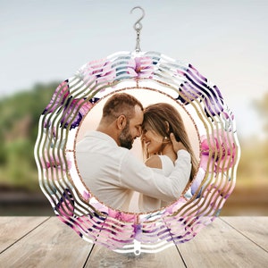 Pode incluir: Um moinho de vento de metal com um design circular que apresenta uma foto de casal no centro. O moinho de vento tem um design em camadas com acentos rosa, roxo e dourado. O fundo é uma imagem desfocada de um campo verde.