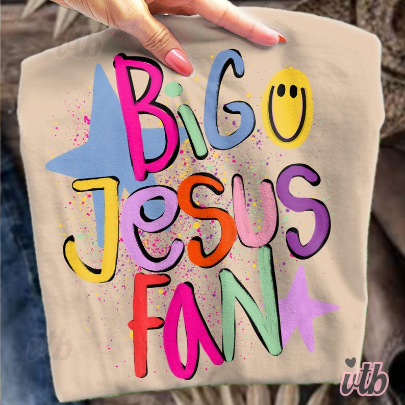 以下が含まれることがあります： ベージュ色のTシャツ。カラフルなバブル文字で「Big Jesus Fan」と書かれています。デザインには星とスマイリーフェイスが含まれています。Tシャツは手で持たれています。
