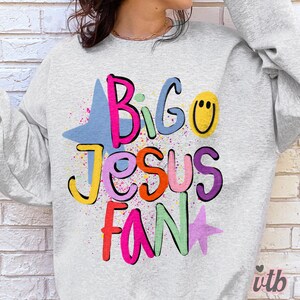 以下が含まれることがあります： 「Big Jesus Fan」の文字がカラフルで遊び心のあるレタリングで描かれた、ライトグレーのスウェットシャツ。デザインには青い星、黄色のスマイリーフェイス、ピンクの星が含まれ、カラフルなペイントスプラッターが施されています。