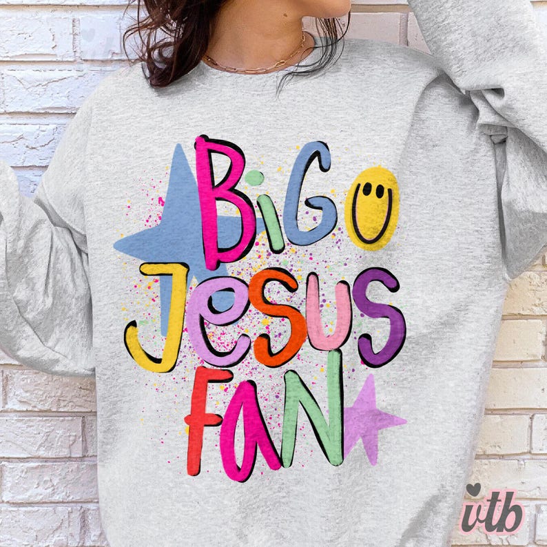 以下が含まれることがあります： 「Big Jesus Fan」の文字がカラフルで遊び心のあるレタリングで描かれた、ライトグレーのスウェットシャツ。デザインには青い星、黄色のスマイリーフェイス、ピンクの星が含まれ、カラフルなペイントスプラッターが施されています。