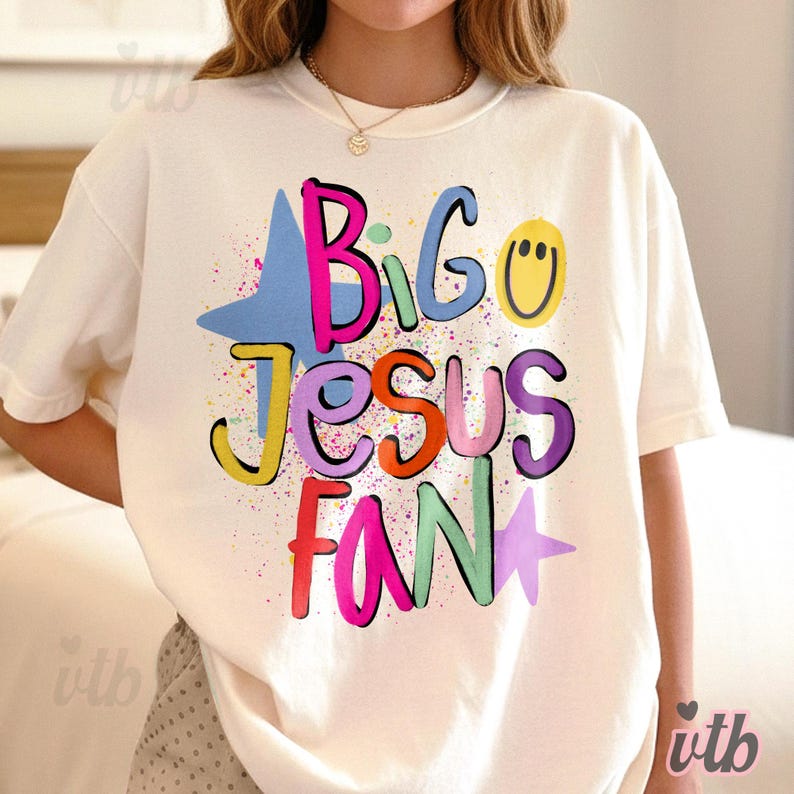 以下が含まれることがあります： 「Big Jesus Fan」の文字がカラフルで遊び心のあるフォントでプリントされた、クリーム色のTシャツ。デザインには星とスマイリーフェイスが含まれ、ペイントスプラッターのアクセントが施されています。カジュアルで快適なTシャツです。