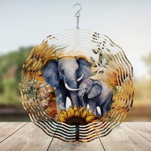Peut inclure: Un mobile de vent en métal avec un design circulaire représentant une mère et son bébé éléphant debout devant un tournesol. L'arrière-plan est un design de style aquarelle de feuilles et de tournesols dans des tons de jaune, orange et vert.
