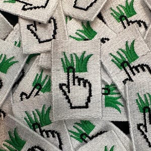 May include: A collection of white embroidered patches. Each patch features a pixelated black hand cursor pointing upwards, above a green grass design. The patches are densely packed together, creating a textured surface.