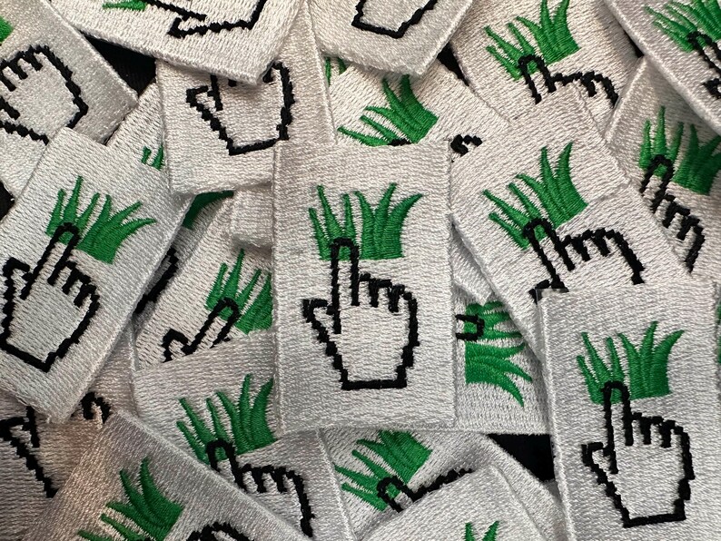 May include: A collection of white embroidered patches. Each patch features a pixelated black hand cursor pointing upwards, above a green grass design. The patches are densely packed together, creating a textured surface.