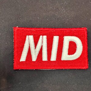 May include: A rectangular red patch with the word "MID" embroidered in white. The patch is likely designed to be sewn or ironed onto clothing or fabric items. The background is a dark, textured surface.
