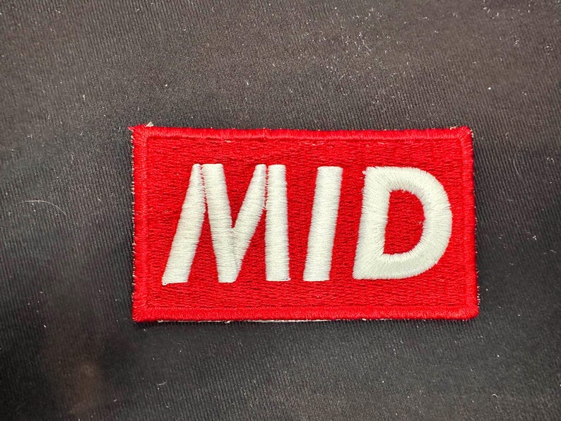 May include: A rectangular red patch with the word "MID" embroidered in white. The patch is likely designed to be sewn or ironed onto clothing or fabric items. The background is a dark, textured surface.