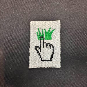 May include: Embroidered patch featuring a pixelated hand cursor pointing upwards towards green grass. The patch is rectangular with a light gray background and black outline. The patch is likely for clothing or accessories.