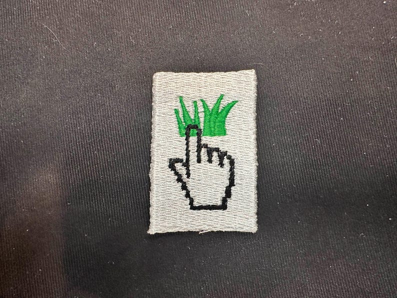 May include: Embroidered patch featuring a pixelated hand cursor pointing upwards towards green grass. The patch is rectangular with a light gray background and black outline. The patch is likely for clothing or accessories.