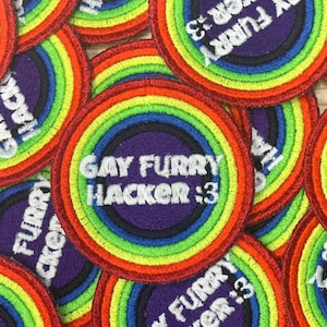 Puede incluir: Una colección de parches bordados circulares. Cada parche presenta un diseño de arcoíris alrededor de un centro púrpura. El texto "GAY FURRY HACKER :3" está bordado en blanco. Los parches son coloridos y vibrantes.