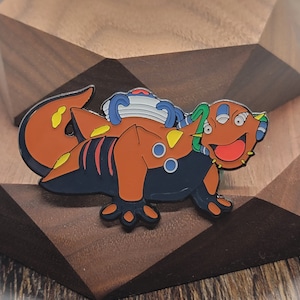 May include: A colorful enamel pin featuring a cartoon creature with a large head, three eyes, and a wide open mouth. The creature is orange, black, and green with blue accents. The pin is on a brown wooden surface.