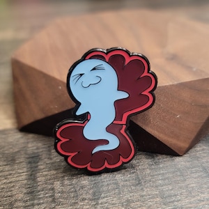 Könnte beinhalten: Ein blauer Geister-Emaille-Pin mit einem lächelnden Gesicht, vor einem dunkelroten, gewellten Hintergrund mit schwarzem Umriss. Der Pin ist ein skurriles Accessoire.