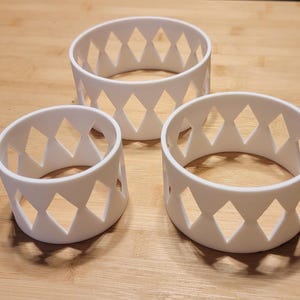 Puede incluir: Tres soportes cilíndricos blancos con recortes en forma de rombo. Los soportes son de diferentes alturas y se encuentran sobre una superficie de madera de color marrón claro. El diseño es simple y moderno.