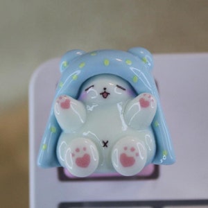 May include: A small, white ceramic figurine of a bear wearing a blue blanket with yellow stars. The bear is sitting up with its paws raised and has pink paw pads. The figurine is on a white keyboard.