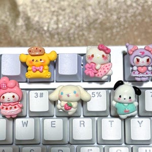 May include: A collection of colorful character keycaps, including a yellow dog with a bow tie, a white cat with a pink bow, and a pink character with a bow. The keycaps are placed on a white keyboard.