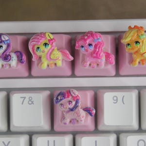 Peut inclure: Cinq touches de clavier en forme de poney de dessin animé colorés. Les touches sont roses, jaunes, violettes et bleues. Les touches sont sur un clavier blanc avec des touches roses.