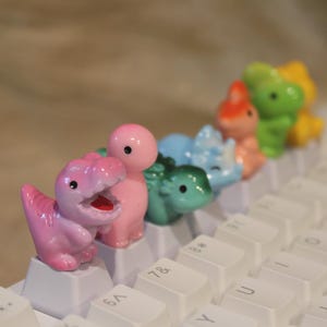 May include: A series of small, colorful dinosaur-shaped keycaps attached to the top of a white computer keyboard. The figures include a pink T-Rex, a pink round dinosaur, a dark green dinosaur, a light blue dinosaur, an orange dinosaur, and a green one.