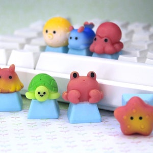 May include: A collection of custom mechanical keyboard keycaps featuring miniature sculpted sea creatures including a yellow pufferfish, blue slug, red octopus, orange crab, green turtle, pink crab, and a yellow and pink starfish on light blue bases.