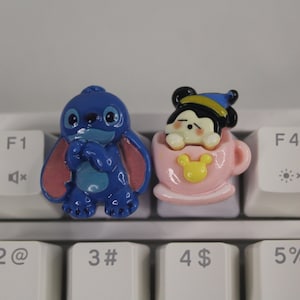 Puede incluir: Dos pequeñas figuras de personajes azules y rosas en un teclado blanco. La figura azul es Stitch de Lilo & Stitch, y la figura rosa es un personaje de Mickey Mouse en una taza de té rosa.