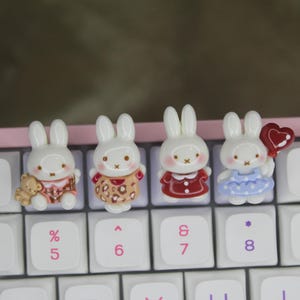Puede incluir: Cuatro pequeñas teclas de teclado blancas con forma de conejo en tres dimensiones están fijadas en un teclado de ordenador blanco con letras rosas. Cada conejo lleva un traje diferente, incluyendo un oso marrón, un vestido de leopardo, uno rojo y uno azul.