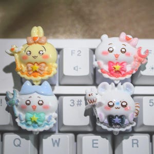 May include: Four colorful, cartoon-style keycaps with cute character designs are arranged on a white keyboard. The keycaps feature characters in yellow, pink, blue, and white, with various accessories and expressions. The keyboard keys are white.
