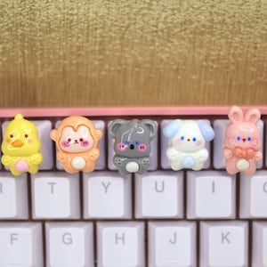 Może przedstawiać: Pięć kolorowych nakładek na klawisze z motywami kreskówek zwierząt. Nakładki są żółte, pomarańczowe, szare, niebieskie i różowe. Nakładki są ułożone w rzędzie na białej klawiaturze.