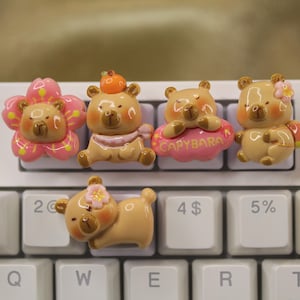 Op de afbeelding: Vijf kleine, bruine, keramische beeldjes van capibara's met verschillende accessoires. De capibara's dragen een roze bloem, een roze sjaal, een roze wolk met het woord "CAPYBARA" erop, een roze bloem en een roze bloem. De beeldjes staan op een wit toetsenbord.