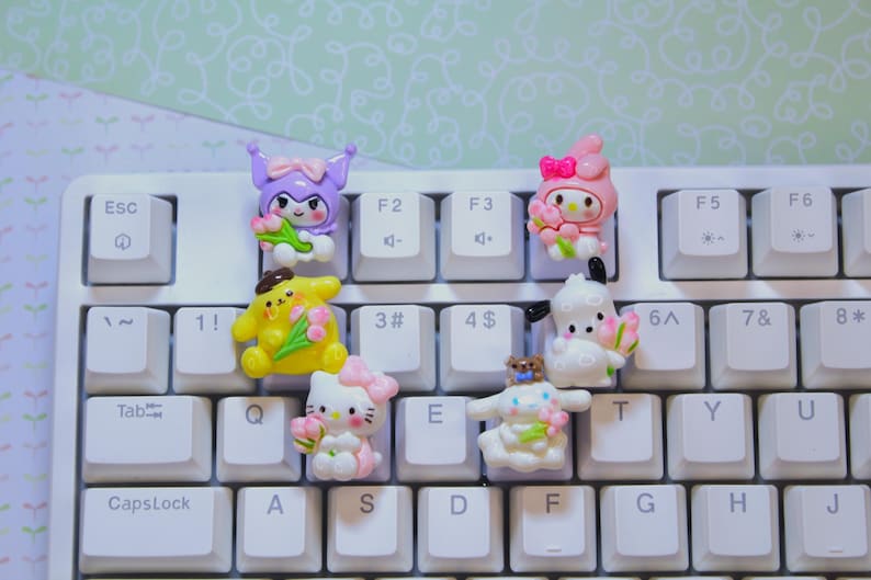 K&ouml;nnte beinhalten: Eine wei&szlig;e Tastatur mit bunten Figuren-Tastenanh&auml;ngern. Die Anh&auml;nger zeigen Comicfiguren, die Blumen halten. Zu den Figuren geh&ouml;ren eine lila Katze, ein gelbes K&uuml;ken, ein wei&szlig;es Kaninchen, ein rosa Kaninchen und ein wei&szlig;er B&auml;r.