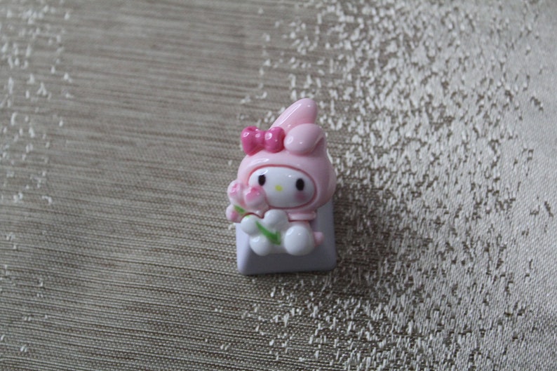 K&ouml;nnte beinhalten: Ein rosa und wei&szlig;er Keycap mit einer Comicfigur mit Hasenohren, einer rosa Schleife und einer Blume. Die Figur h&auml;lt eine Blume in der Hand.