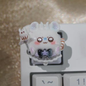 May include: A white, cartoon-style keycap featuring a cute mouse character with large eyes, a bow, and a small creature on its back. The keycap is on a white keyboard, with the "~" and "1!" keys visible.