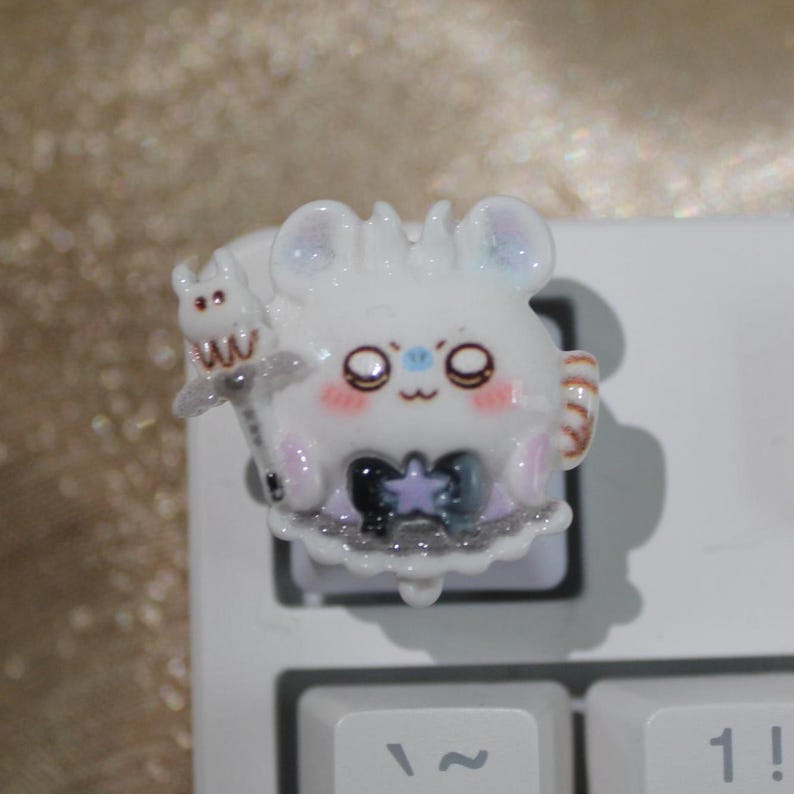 May include: A white, cartoon-style keycap featuring a cute mouse character with large eyes, a bow, and a small creature on its back. The keycap is on a white keyboard, with the "~" and "1!" keys visible.