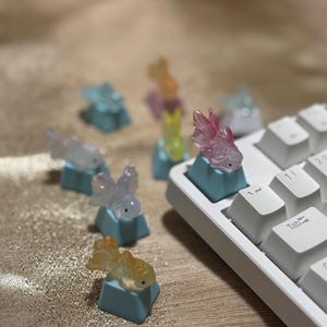 Puede incluir: Teclas de teclado personalizadas con diseños de peces dorados translúcidos y coloridos. Las teclas están montadas en un teclado blanco. Los peces dorados son rosas, amarillos y azules. Las teclas son de color azul claro.