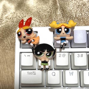 May include: Three Powerpuff Girls character keycaps in Blossom, Bubbles, and Buttercup designs. The keycaps are colorful and detailed, with each character's signature look. They are placed on a white keyboard.