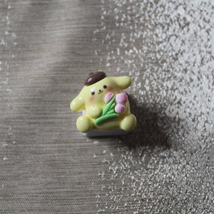 K&ouml;nnte beinhalten: Ein gelber und wei&szlig;er Keycap mit einer Comicfigur, die einen Strau&szlig; rosa Blumen h&auml;lt. Die Figur tr&auml;gt einen braunen Hut.